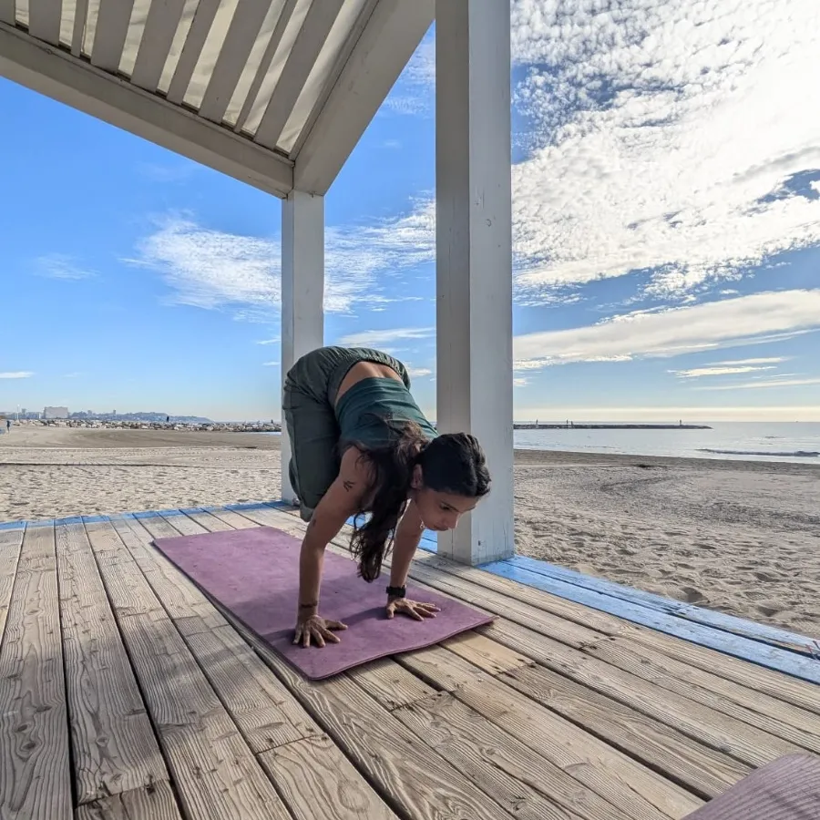 Profesora de yoga entrando en la postura del cuervo sobre una esterilla en una pasarela de playa, con la arena y el mar detrás